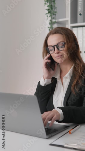 Focused female entrepreneur in formal attire communicates on phone while using laptop in bright office setting. Productivity, leadership, and remote business management concept for corporate success