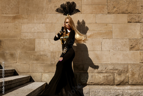 Fotografie Majestic female model in avant-garde flowing black gown with metallic gold armor and elaborate feather headdress posing against a textured stone wall in a high-fashion fantasy editorial