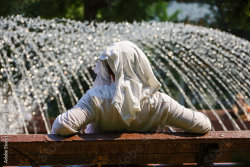 Man with covered head sitting on fountain background in city park. Hot sunny weather