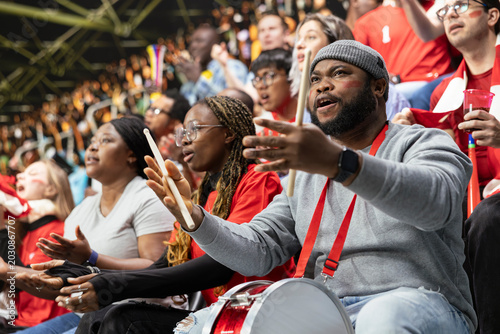 Tablou pe pânză Black football supporter with drum gesturing anxiously with open hands in red stadium crowd during tense live soccer match
