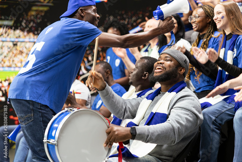 Quadro su tela Black football capo within blue supporter crowd and directing stadium chants with drum leader during intense soccer match