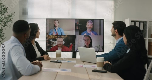 Multicultural office team joins video meeting with remote coworkers on large screen in boardroom. Colleagues watch display and discuss project updates together