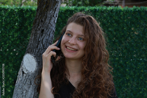 Young woman smiles while talking on the phone near a tree at a park on a sunny day in the afternoon