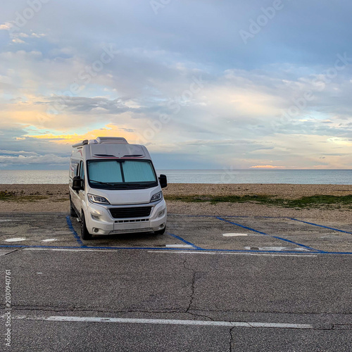 Foto A white modern recreational vehicle camper van parked in a designated parking lot next to a pebble beach, looking out over a calm sea under a beautiful sunset sky