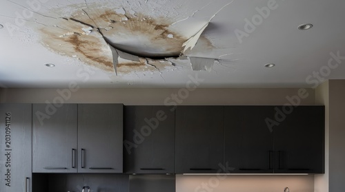 A kitchen ceiling shows severe water damage with peeling paint and a large brown stain above dark cabinetry.