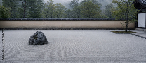 A serene Japanese zen garden features a prominent rock surrounded by meticulously raked gravel, evoking a sense of tranquility amidst lush greenery and a gray sky.