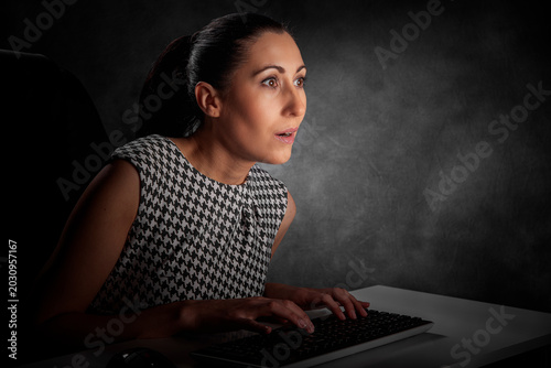 Fotografie A surprised businesswoman sits at her desk at night, staring in shock at her glowing computer screen