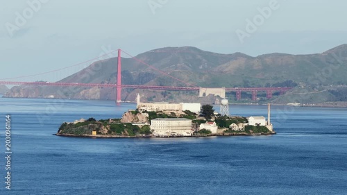 Alcatraz Island in San Francisco Bay with historic prison buildings and lighthouse with Golden Gate Bridge and Marin Headlands in background under clear blue sky at daytime. Aerial wide shot.