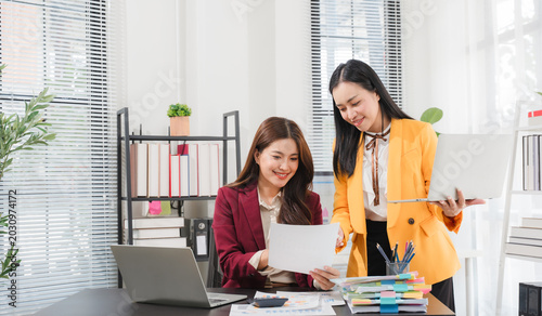 Photos Collaborative Workspace: Two professional women engage in a productive discussion in a modern office setting, reviewing documents and laptops, showcasing teamwork and business acumen