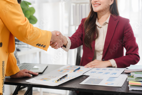 Successful Business Partnership: Two business partners shake hands over a desk laden with documents and charts, sealing a deal with a warm smile, symbolizing trust and collaboration.