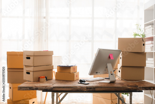 Moving Day Ready: Cardboard boxes are stacked high on a rustic wooden desk in an office setting, hinting at relocation or expansion. A computer monitor sits ready for the next chapter.