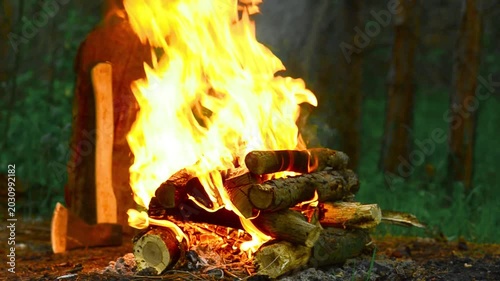 Close up of burning bonfire with tourist ax stuck in chopped wood log nearby, cinematic campfire with bright orange flames, flying sparks and smoke in green forest camping site background in 4K