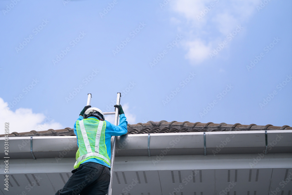Naklejka premium Worker in Safety Vest Climbing Ladder to Inspect Roof Gutter on Residential House Against Blue Sky