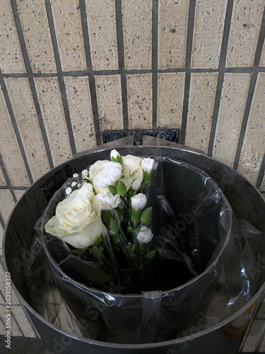 White roses and delicate carnations discarded in a public trash bin against a tiled wall, creating a melancholic urban scene symbolizing loss, rejection, or the fleeting beauty of flowers.