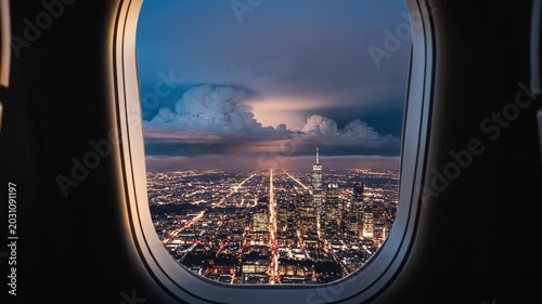 Stunning aerial view of a glowing city skyline at night seen through an airplane window, with dramatic storm clouds and lightning in the background