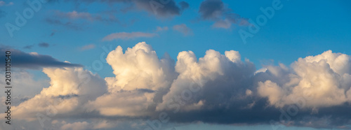 Dramatic cumulus clouds with golden backlighting on blue sky, atmospheric panoramic celestial landscape in evening light