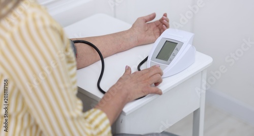 Senior woman measuring blood pressure at white table indoors, selective focus