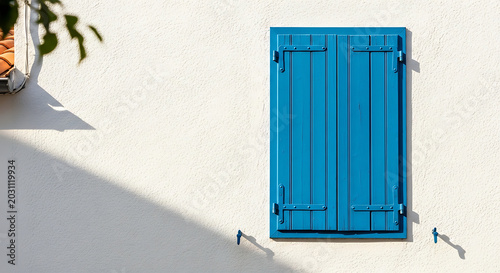 Closed bright blue wooden window shutters on a white textured wall of a Mediterranean house with sunlight and shadows