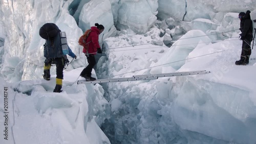 Alpinists with backpacks navigating a dangerous ice crevasse using a ladder bridge in the Himalayas