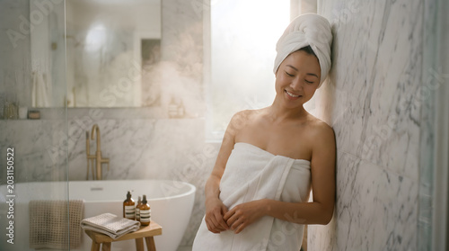 Smiling woman wrapped in white towel after shower standing in marble luxury bathroom