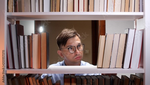 Fotografie Man in eyeglasses looking intently between books on a white bookshelf, carefully