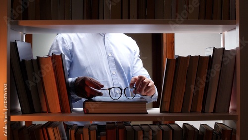 Schilderij op canvas Person stands at a library bookshelf selecting a book while holding reading glas