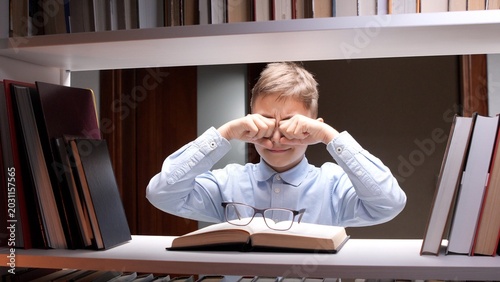 Foto Young boy in a library rubbing dry eyes and grimacing while studying a book, sho