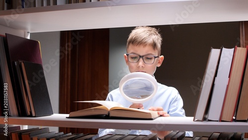 Tablou pe pânză Young boy in glasses reading an open book on a brightly lit shelf using a magnif