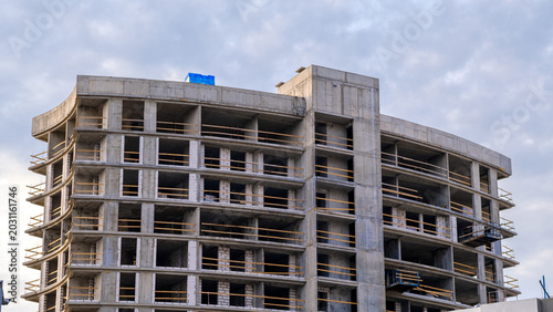 Concrete structure rises against cloudy sky. Windows frame empty spaces for future lives. Construction workers move through unfinished floors.