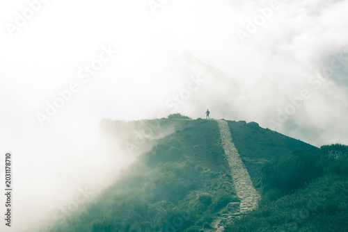 A stone hiking trail leading along a misty mountain ridge toward a lone hiker walking into dense white clouds.
