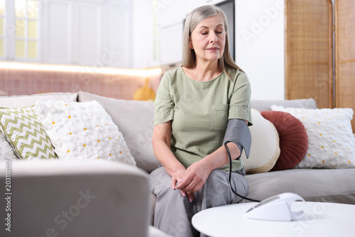 Senior woman measuring blood pressure on sofa at white table indoors