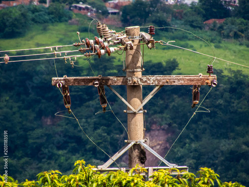 Detailed shot of an electrical distribution pole featuring weathered wooden crossarms, insulators, and power lines against a blurred natural green landscape in rural Brazil.