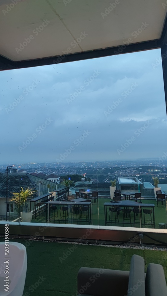 Fototapeta premium View from a modern rooftop cafe terrace overlooking a vast city skyline under a dramatic overcast evening sky.