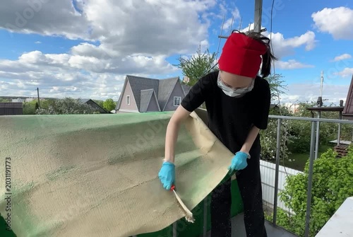 Person cleaning a carpet on a balcony with a metal brush, wearing a mask and goggles, with white debris falling, suitable for cleaning service advertisements, DIY cleaning guides