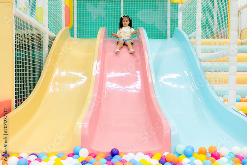 Bilde på lerret Little Girl Sliding into a Colorful Ball Pit in an Indoor Playground,A cheerful little Asian girl with dark hair tied up, wearing a light-yellow t-shirt and light blue shorts, is captured in motion