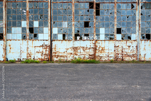 The exterior wall of a desolate, large metal airplane hangar building with half a wall of glass windows. Some glass panes are broken, have holes from rocks or are empty. There are rust patches on the 