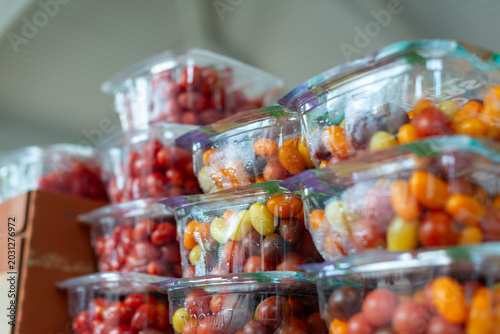 A stack of small, clear plastic containers of raw organic cherry tomatoes on a grocery store shelf in a store for sale. The ripe Roma tomatoes are firm with thick, shiny skin in a square package.  