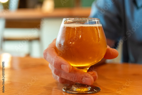 A Caucasian man wearing a blue shirt holds a pale ale beer in a clear, tulip-shaped glass on a wooden table. The glass is filled with a cold golden craft drink with foam on top of the pint glass.