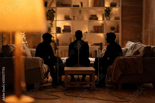 Siluetas de tres hombres grabando un podcast nocturno en una sala acogedora con micrófonos de pie. Concepto de entrevista, programa de radio digital y conversación íntima