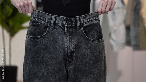 Slim woman lifts loose old black jeans to waist and lets them fall, falling at home. Concept of weight loss result, healthy lifestyle, wellness, body transformation, fitness progress and size change