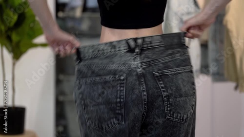 Rear, back view of slim woman in old wide jeans after weight loss, joyfully swaying hips in home interior. Concept of healthy lifestyle, body transformation, diet progress and clothing size change
