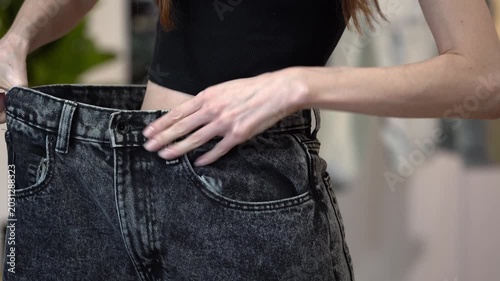 Slim woman fitting old big jeans and pulling waistband wide after weight loss, home interior background. Concept of healthy diet, proper nutrition, wellness, slimming success and clothing size change