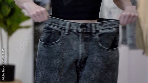 Young slim woman stretches old loose jeans side to side at waist in home interior, showing figure change after weight loss. Concept of slimming success, body transformation and wellness progress