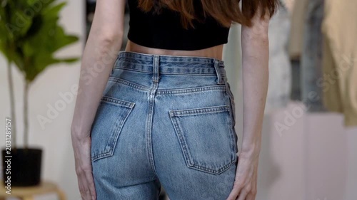 Young Caucasian woman in fitted blue high-waisted jeans turns gracefully and strokes hips in wardrobe home interior. Concept of flattering denim fit, feminine confidence, slim waist and casual style