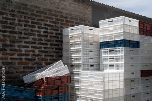 Stacks of empty white and colored plastic crates outdoors against a brick wall under a blue sky. Eye-level view. Concept of agricultural storage, logistics, and supply chain