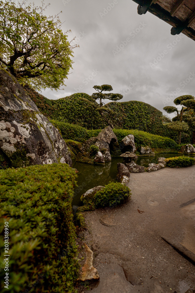 Obraz premium Chiran, Japan - March 25, 2026: Traditional Japanese garden with reflective wet ground and sculpted shrubs in Chiran Samurai Village, Kagoshima, Japan. 