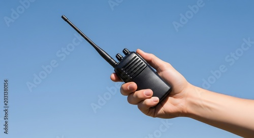 A hand holding a black walkie talkie radio against a clear blue sky