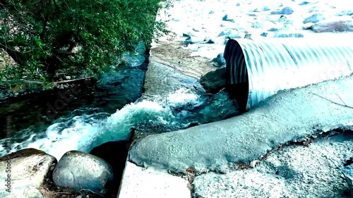 Water rushes from a large metal pipe into a rocky stream below with white foam and concrete edges surrounded by stones and green foliage in a natural outdoor setting
