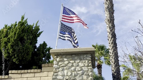 Two American flags wave on a stone pillar with green trees palm trunks and a blue sky in the background on a sunny day in an outdoor setting
