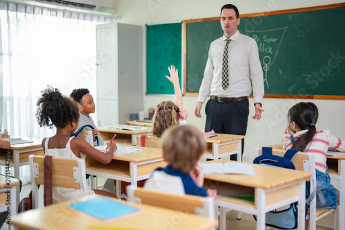 Elementary school teacher leading classroom discussion while students raise hands during interactive lesson in bright multicultural learning environment.